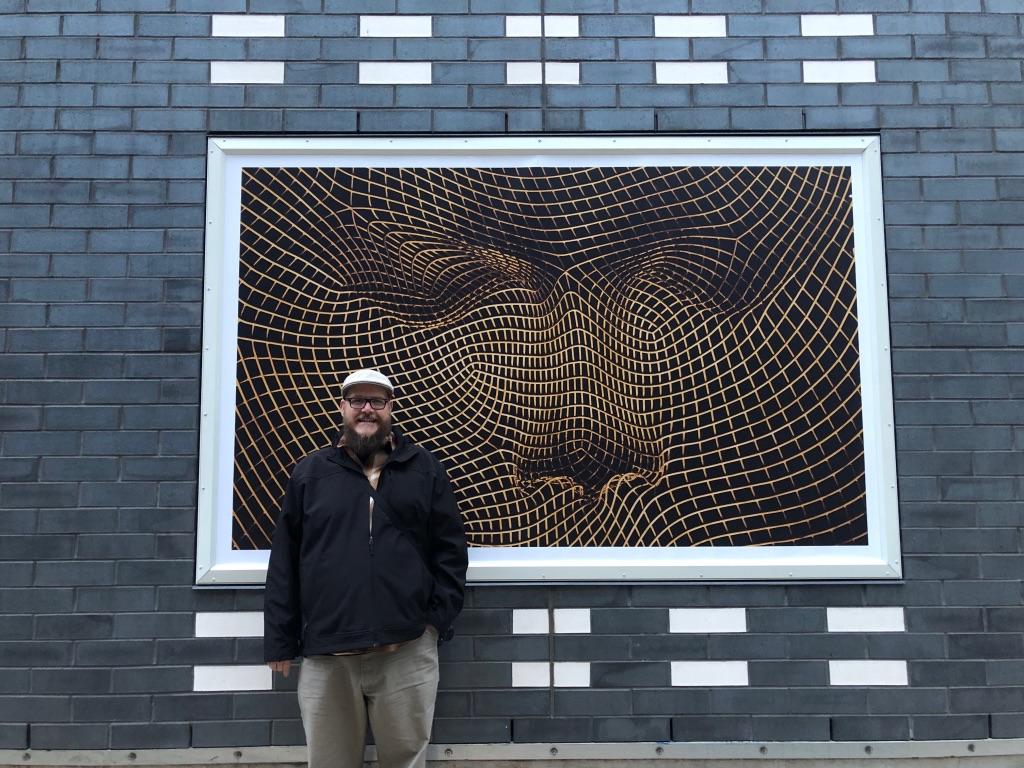 i.m. ruzz standing in front of one of his large exterior photographs installed at Alt Hotel Calgary.