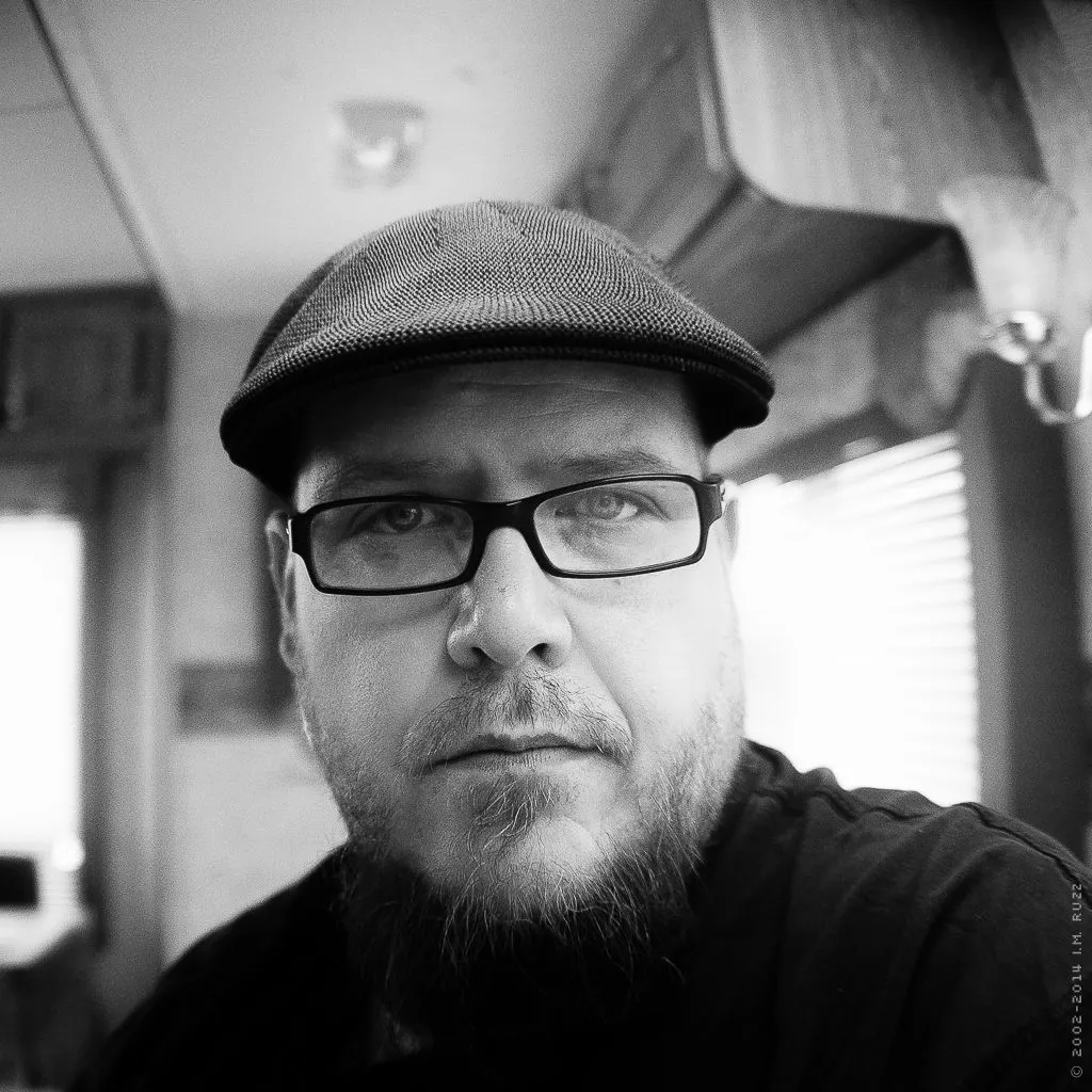 Black-and-white self-portrait of i.m. ruzz wearing a flat cap and glasses.